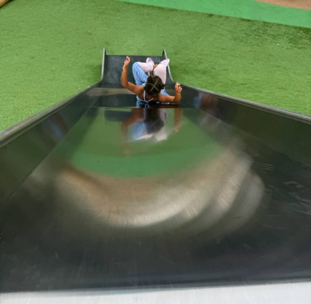 Embankment slide installation on a turf slope, with a child riding a wide stainless steel flat-bed slide
